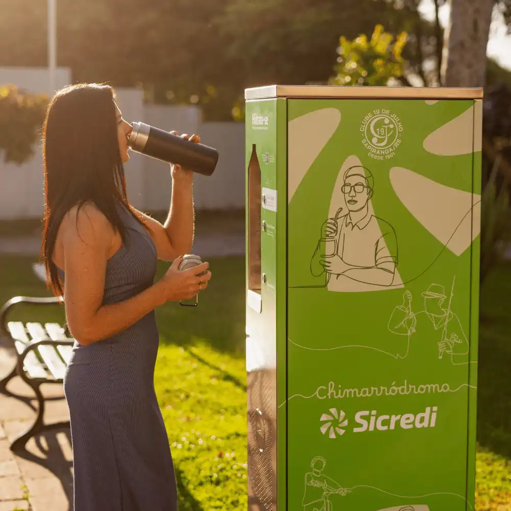 Mulher bebendo da garrafa ao lado de uma estação Sicredi